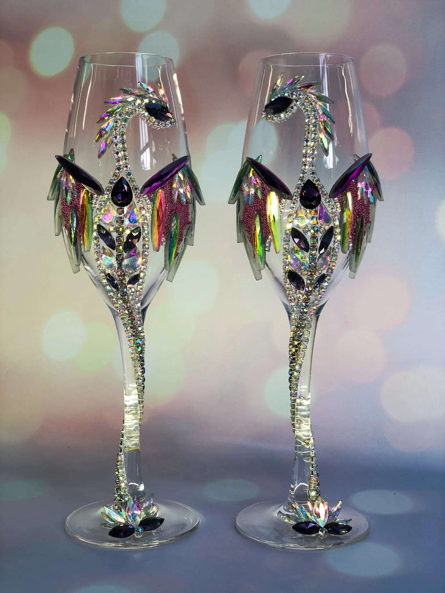 Pair of crystal-clear wineglasses adorned with vibrant holographic dragon motifs; the gems sparkle against a backdrop of soft pastel bokeh lights.