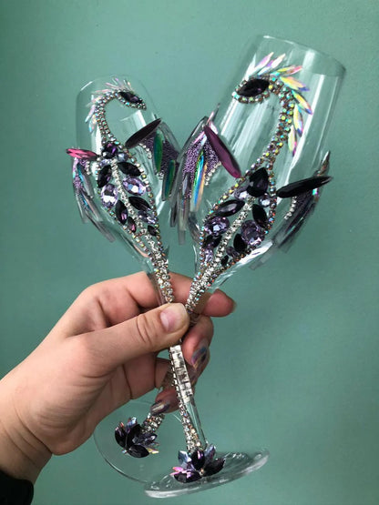 Hand gracefully holding two bedazzled wineglasses by their shimmering, gem-encrusted stems, emphasizing their elegance and the fine craftsmanship; juxtaposed against a cool mint-green background